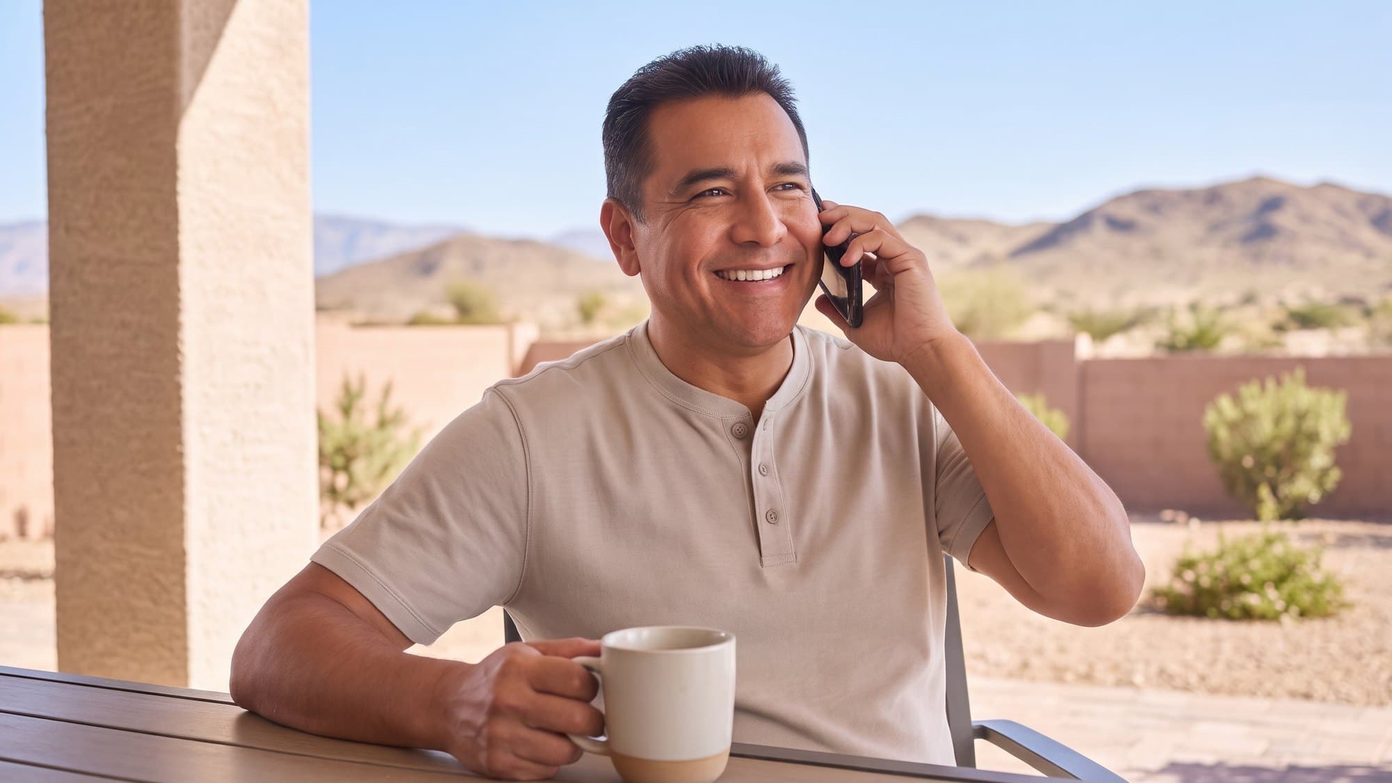 A smiling man talking on the phone with a coffee mug on a sunny home patio