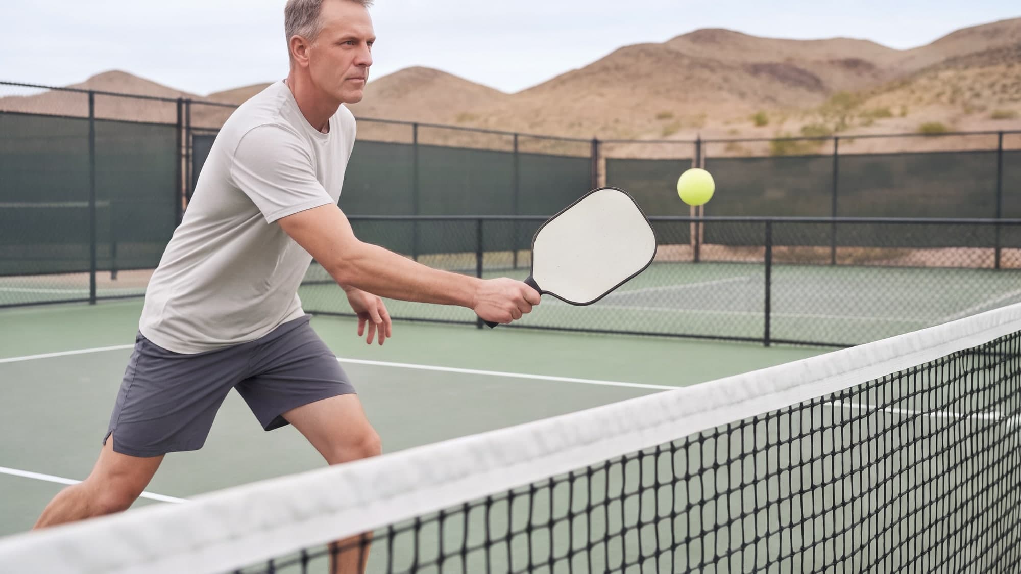 A man at the kitchen line of a pickleball court in Henderson, mid-volley with a plain paddle