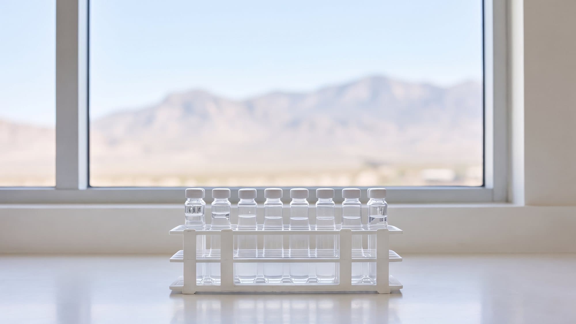 A row of small sealed glass vials in a white rack on a bright bench in front of a window with mountains beyond