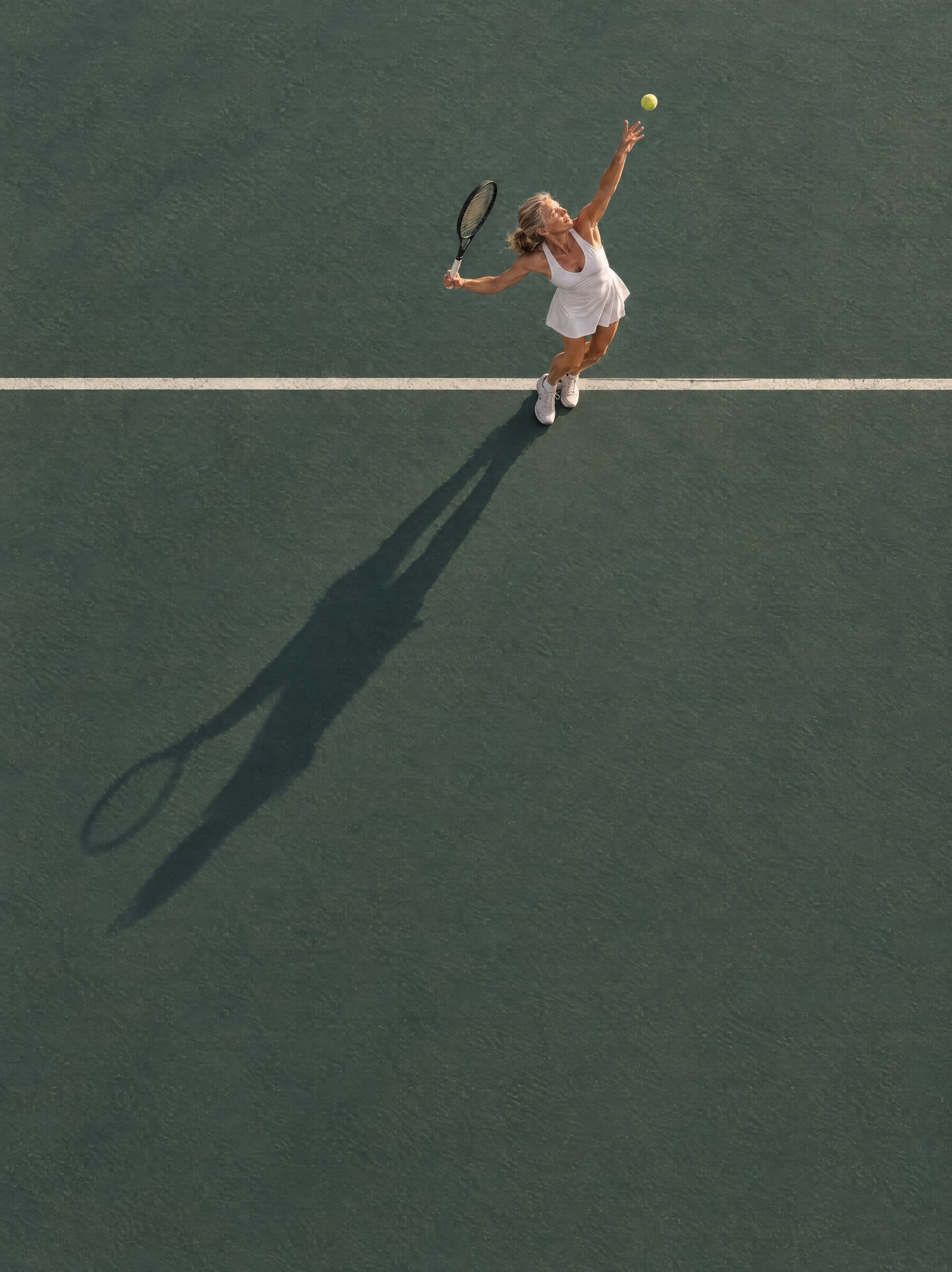 A woman serving on a tennis court, seen from above, her long shadow across the court