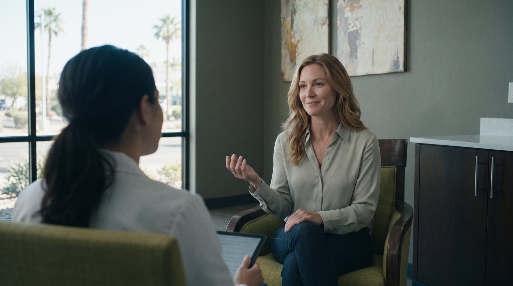A woman in a sage blouse talks with her nurse practitioner in the consultation room at GIOSTAR Henderson, palm trees in the window behind her