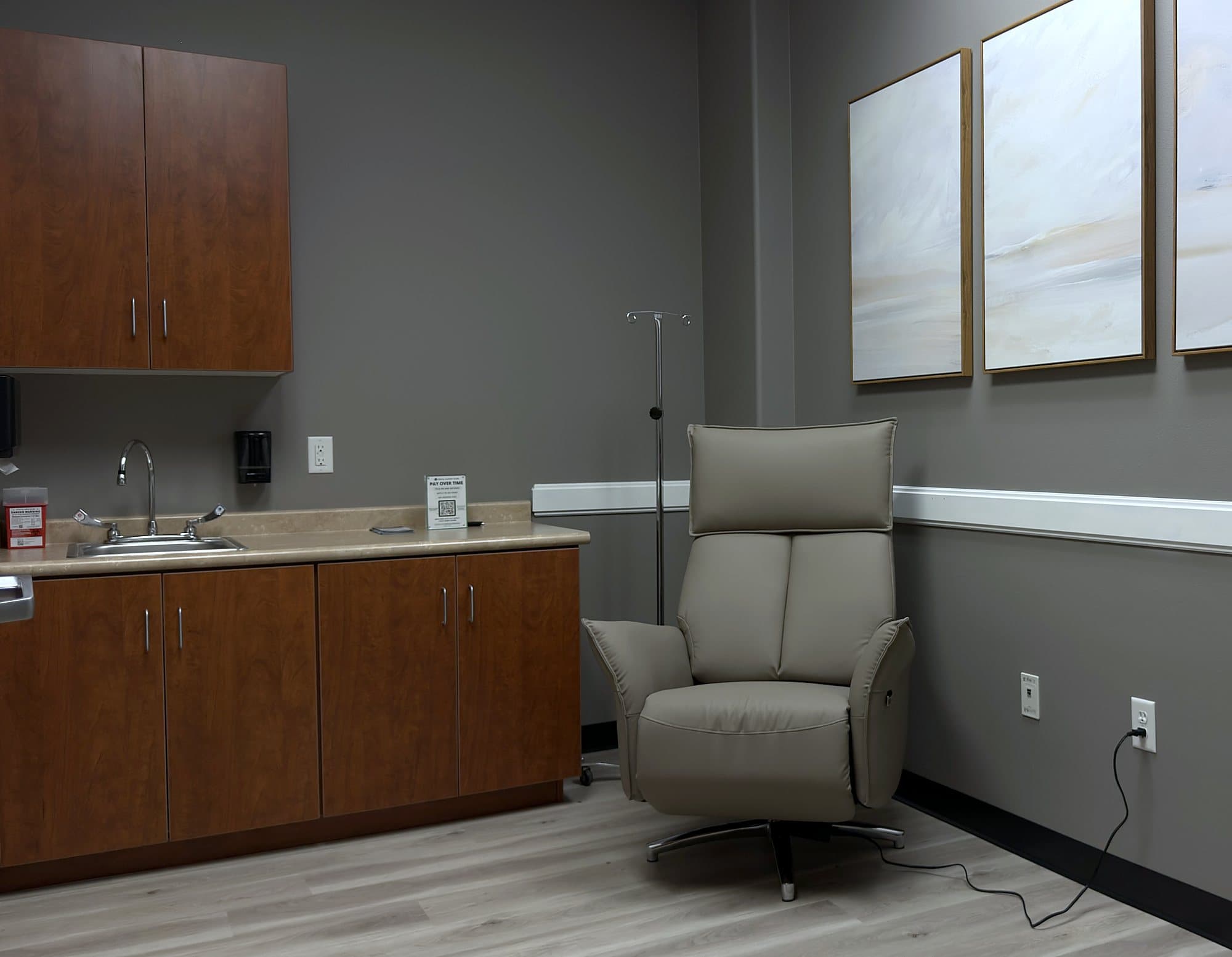 A private IV room at GIOSTAR Henderson with a gray treatment recliner, cabinets and three soft landscape paintings