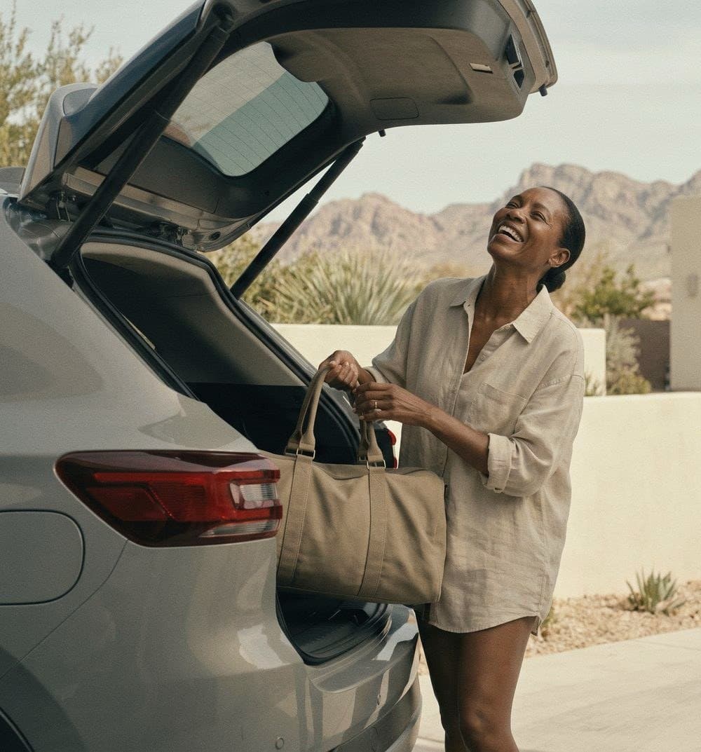 A woman laughs as she lifts a tan duffel bag from the open trunk of a car, desert mountains behind her