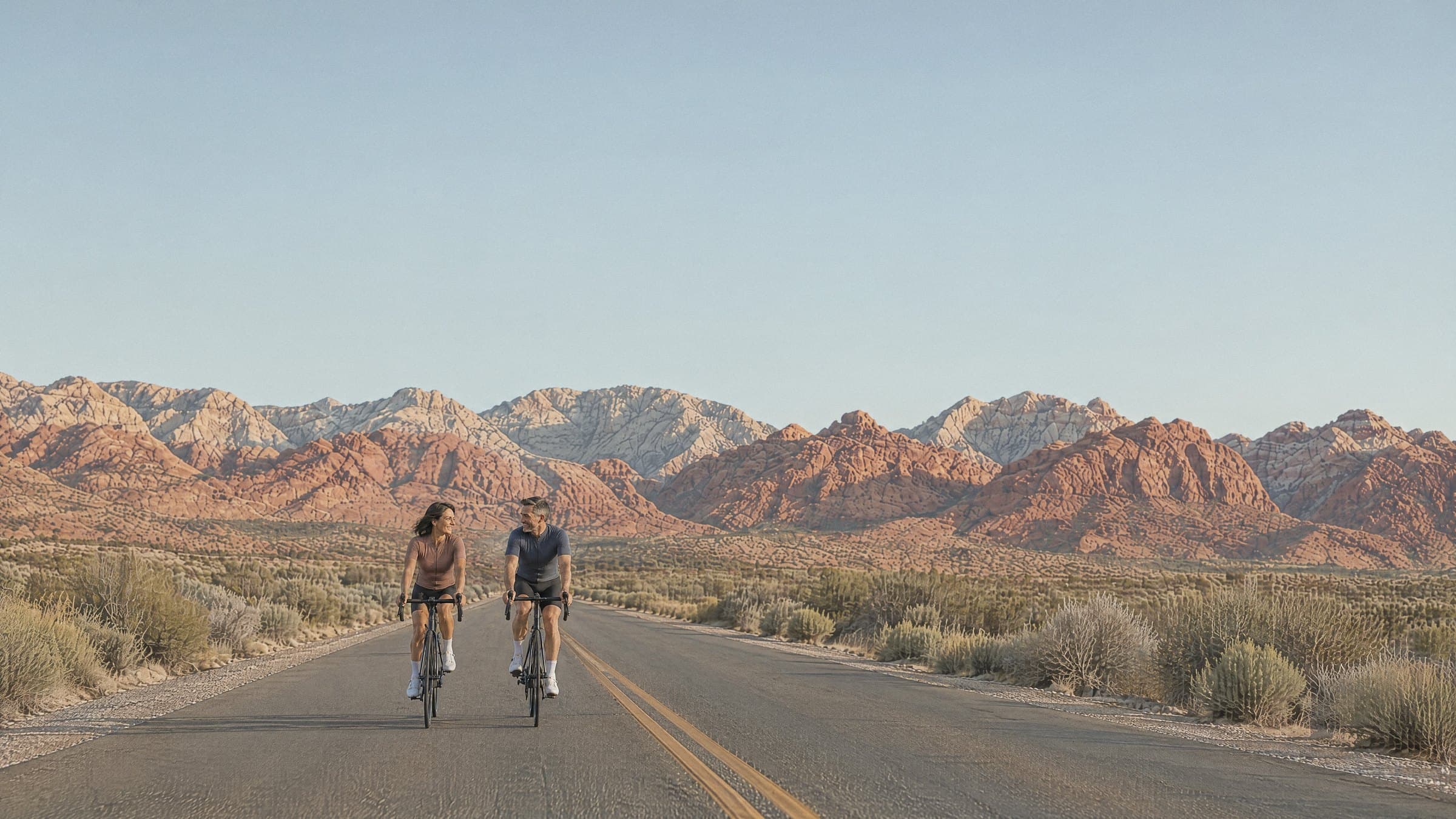 A couple cycling side by side on an open desert road below the red and white cliffs of Red Rock Canyon