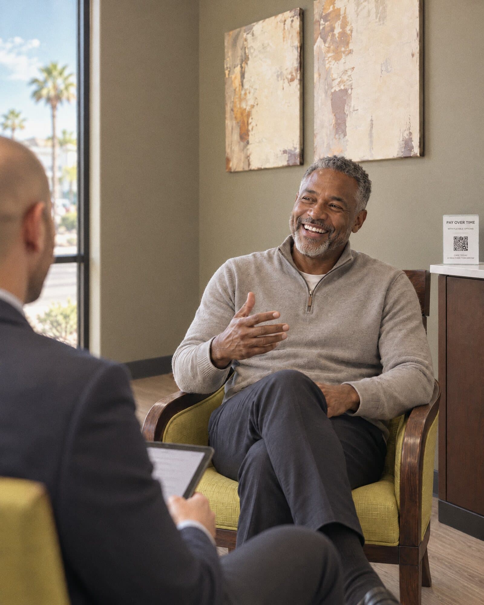 A man in a grey quarter-zip smiling during a consultation in a room at a Henderson wellness center
