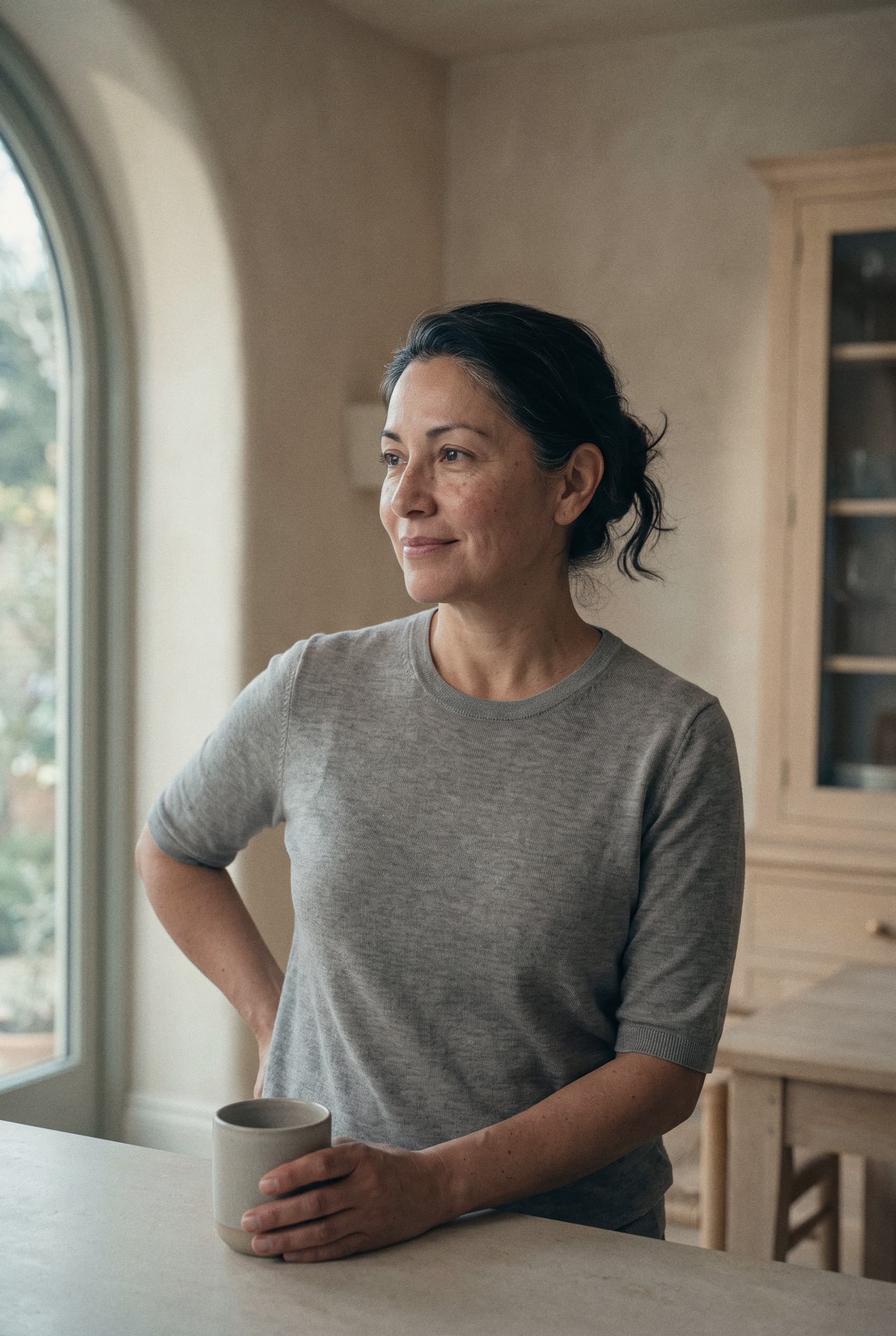 A woman in her fifties holding a ceramic mug and looking out an arched window in a bright kitchen