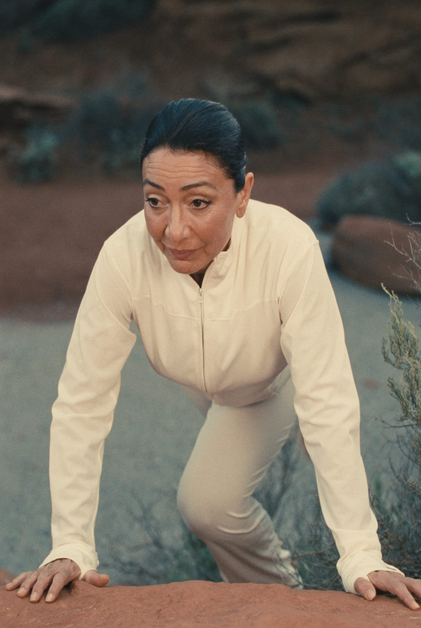 A woman in cream athletic wear climbing a red rock ledge on a desert trail