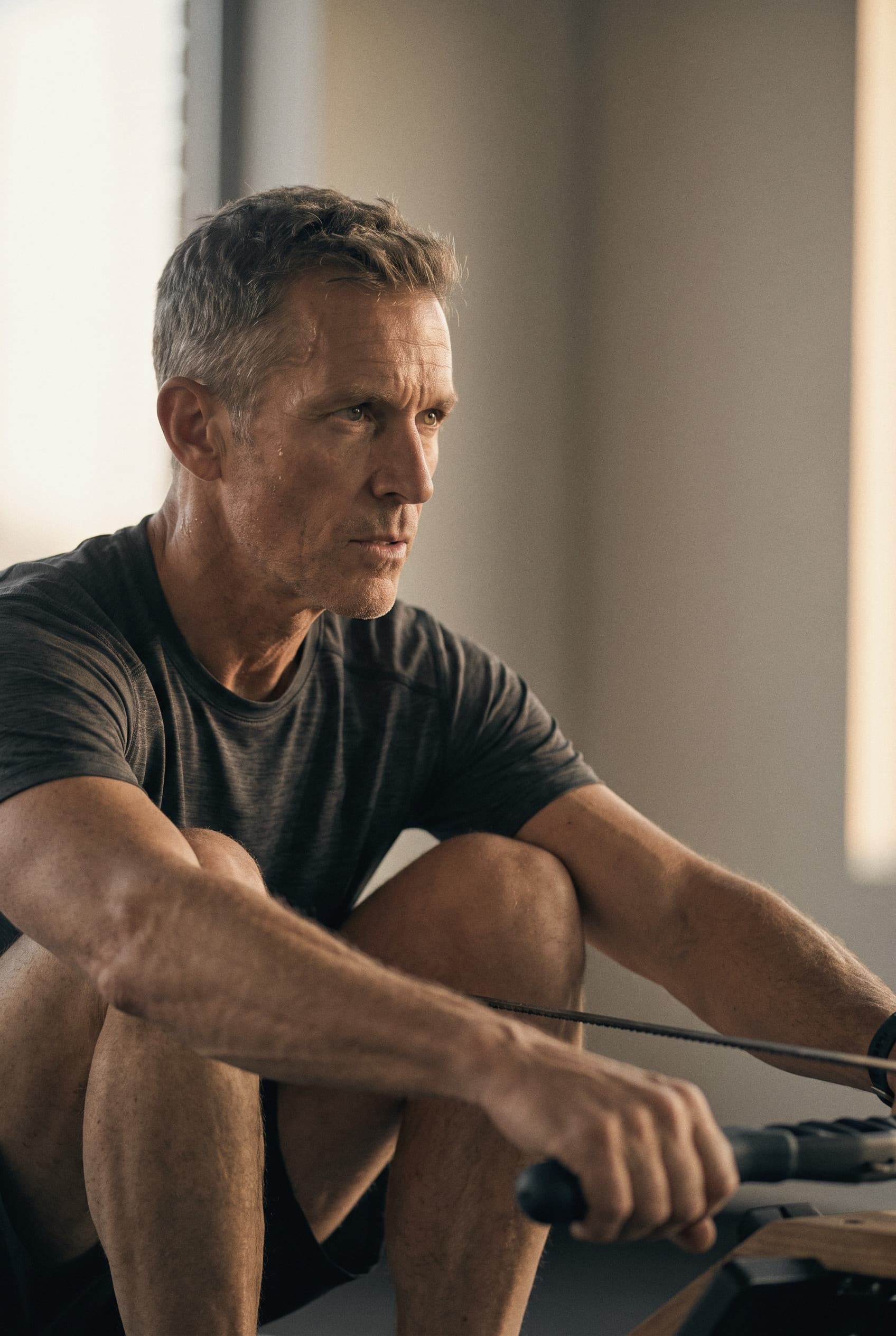 A man in his fifties resting his arms on his knees at a rowing machine, looking ahead in soft window light